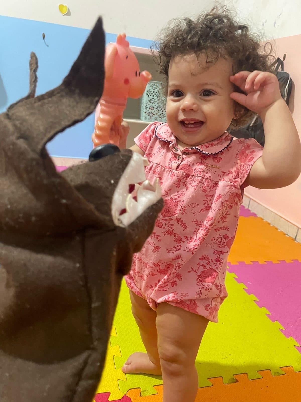 Menina sorrindo de pé no tapete colorido, diante de um fantoche de lobo