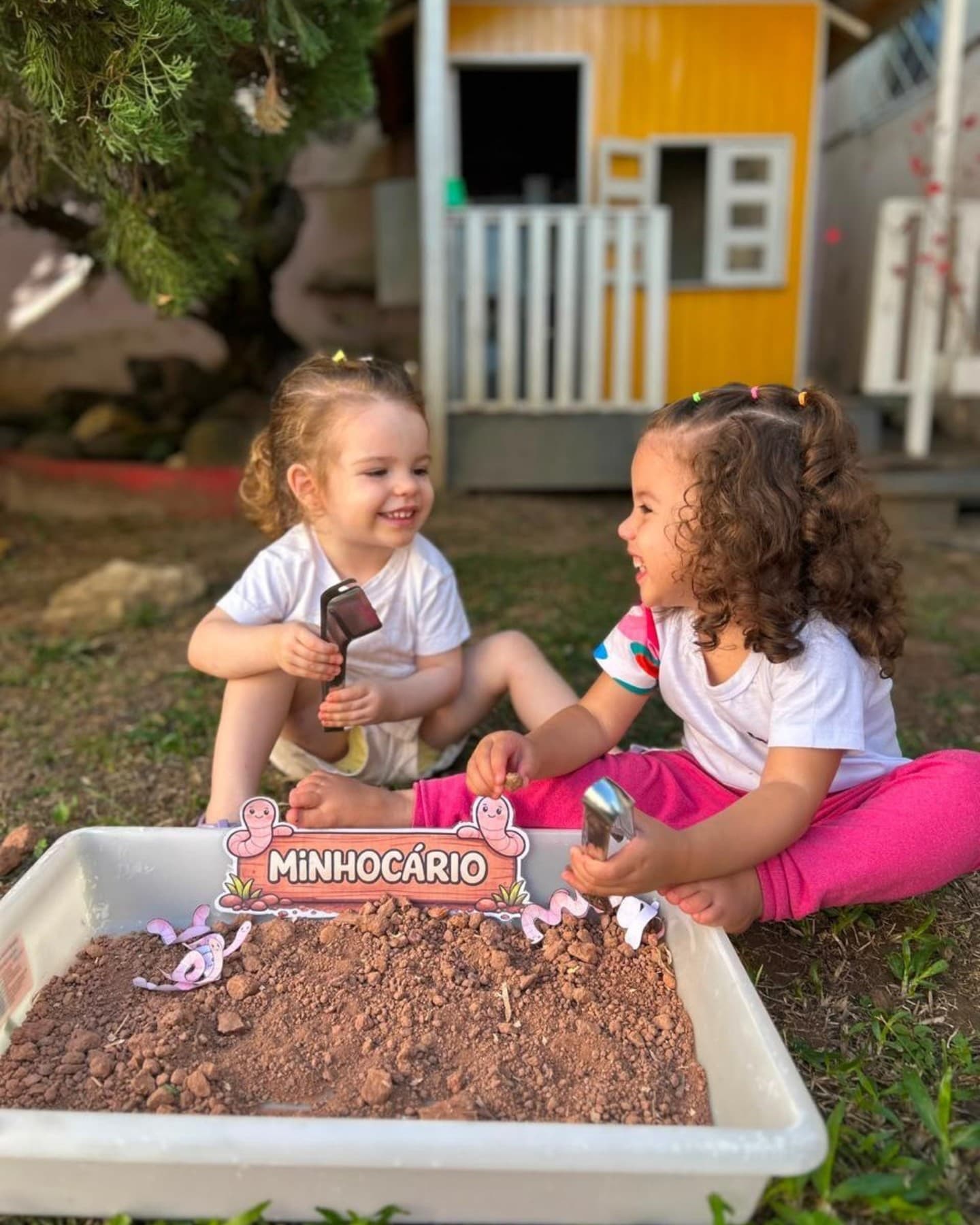 Duas meninas sentadas na grama rindo, com um minhocário de terra na frente