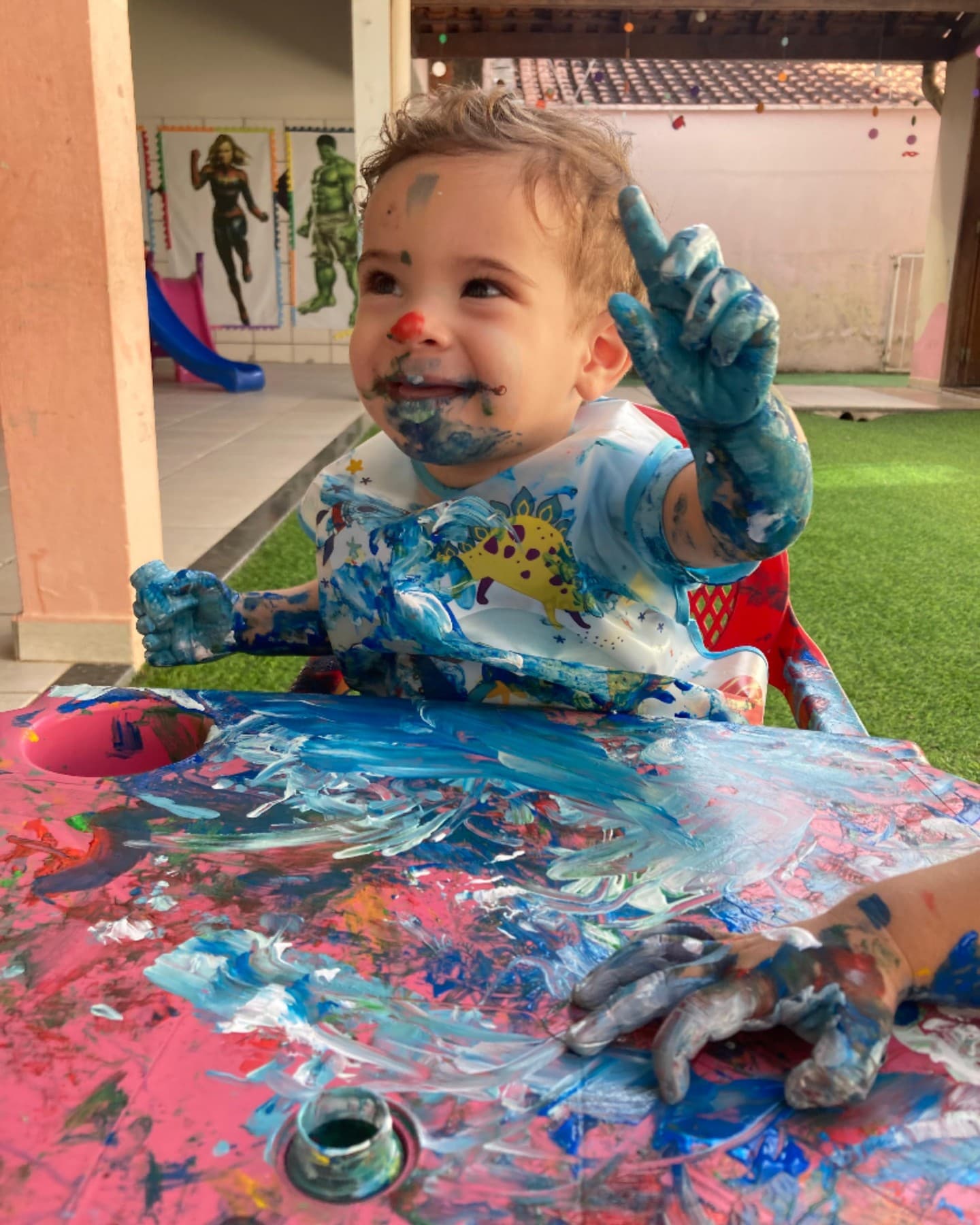 Bebê sorrindo, com as mãos e o rosto cobertos de tinta azul, pintando uma mesa no quintal