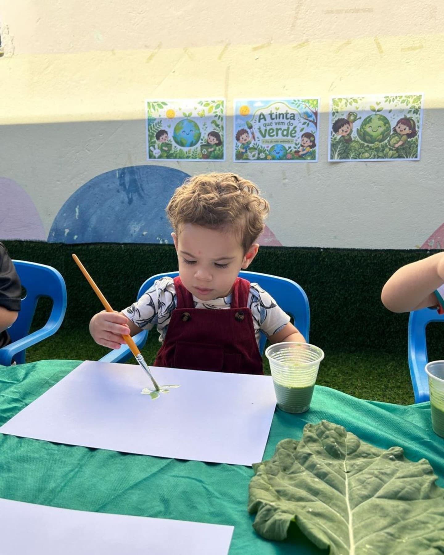 Menino pintando com pincel e tinta verde, com uma folha de couve na mesa